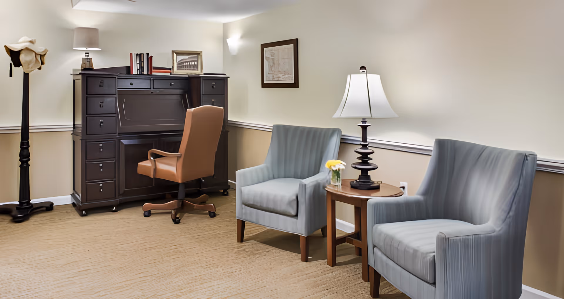 A cozy interior room featuring two light blue upholstered armchairs separated by a wooden side table with a lamp and a small vase of yellow flowers. To the left, there is a dark wooden roll-top desk with a brown office chair on wheels. A tall black coat rack with a hat is positioned near the desk. The walls are painted in a light cream color with a chair rail molding, and there is framed artwork hanging on the wall.