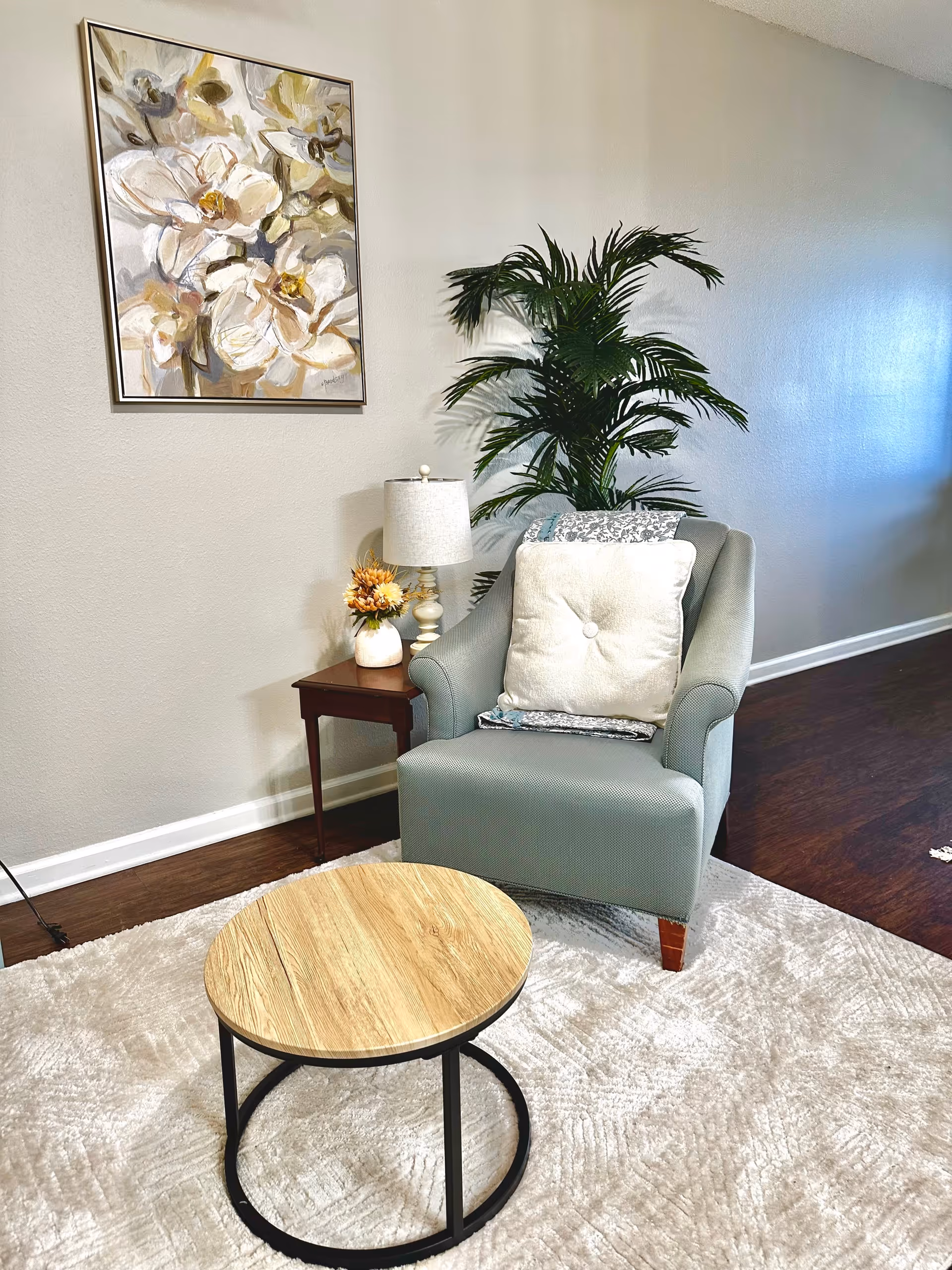 A cozy living room corner featuring a light blue armchair with a decorative pillow, a wooden coffee table, a lamp, and a potted plant, alongside a floral painting on the wall.