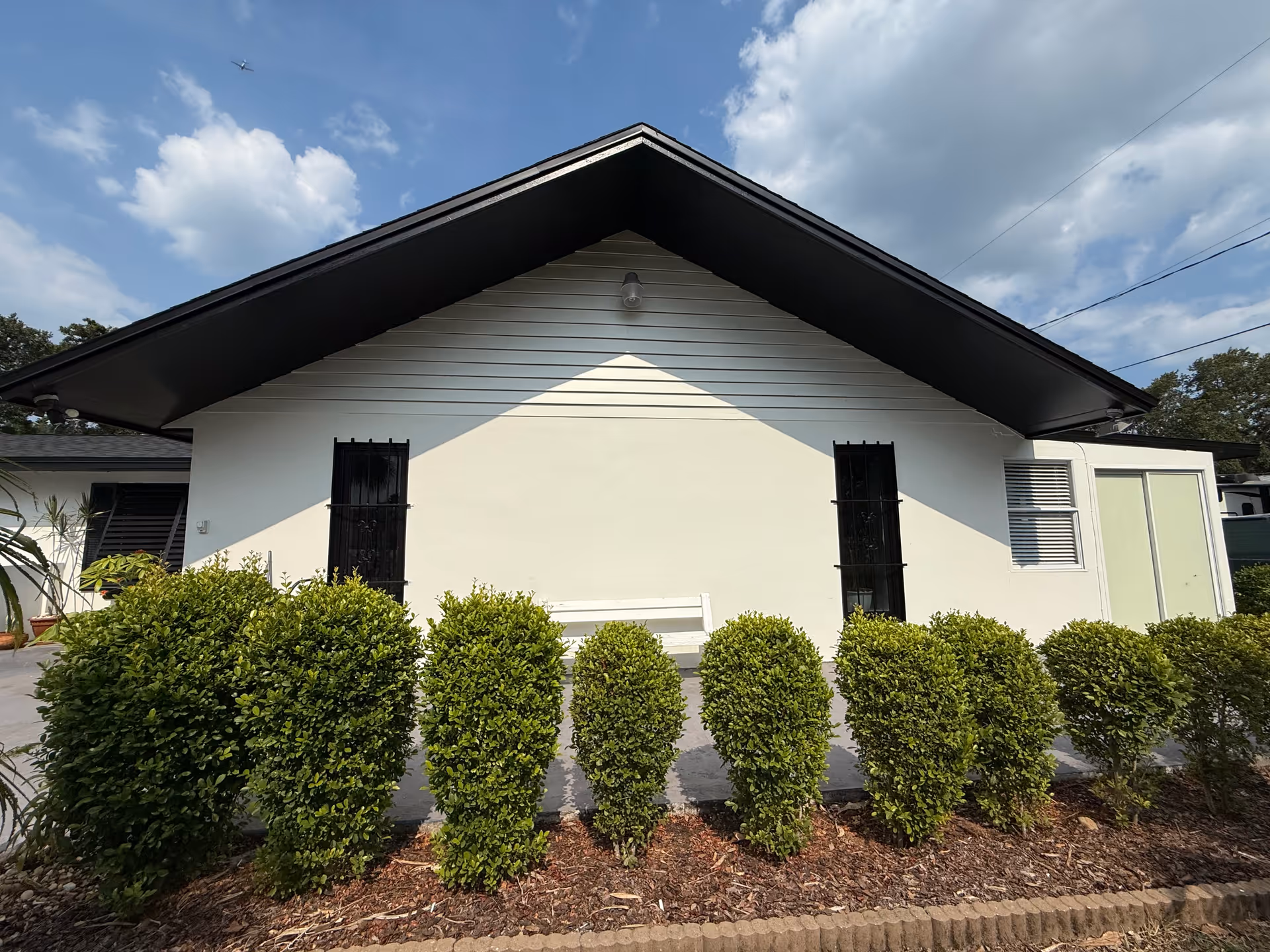Exterior view of a single-story building with a triangular roof and white walls. There are two black barred windows and a white bench between them. In front of the building, there is a row of neatly trimmed green bushes. The sky is partly cloudy.