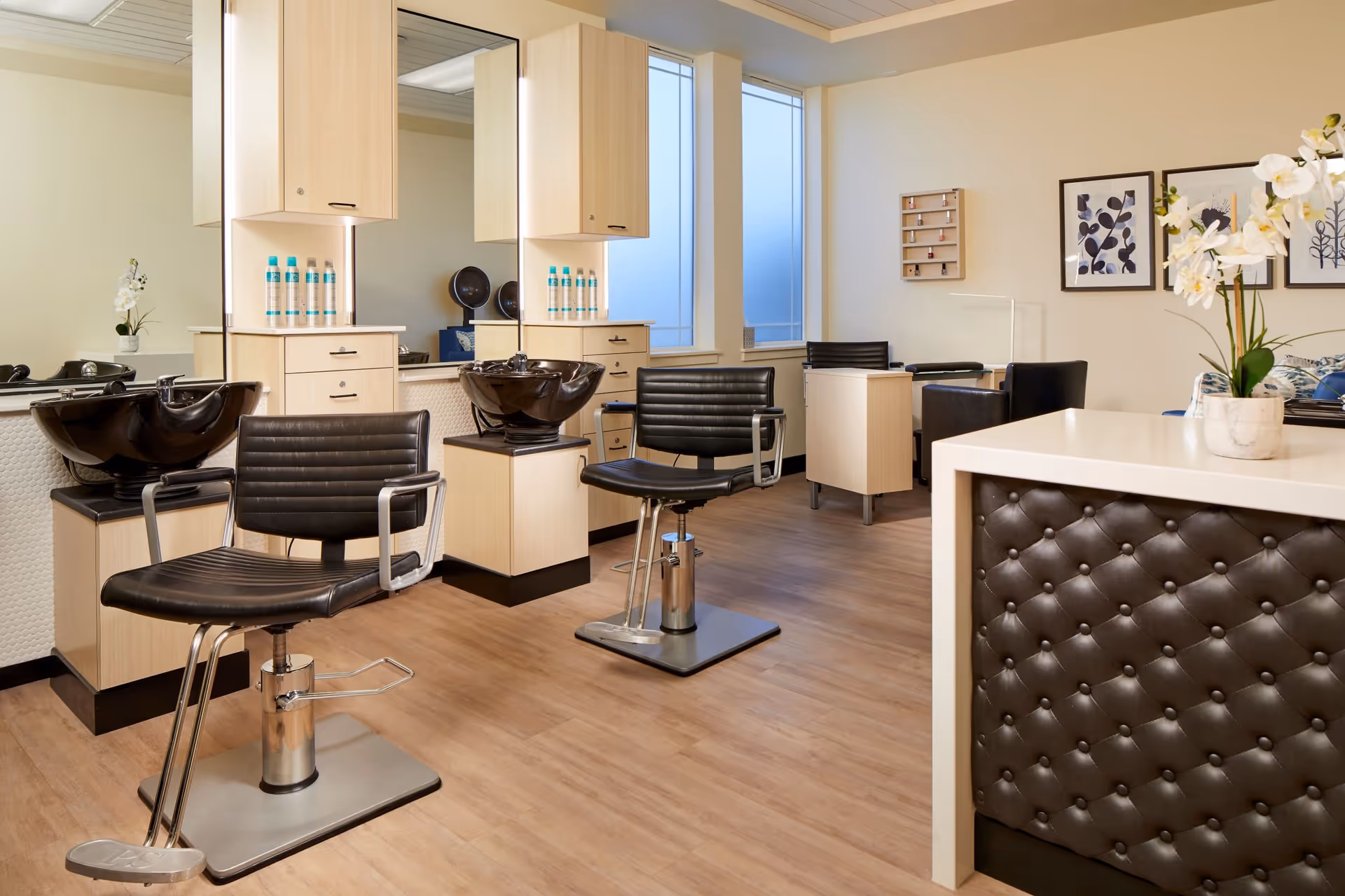 Interior view of a modern salon area with two black salon chairs in front of hair washing stations with black sinks. Light wood cabinetry and large mirrors are behind the chairs. The floor is light wood, and there is a white counter with a tufted dark brown front panel and a white orchid plant on top. The room has large windows and framed botanical artwork on the wall.