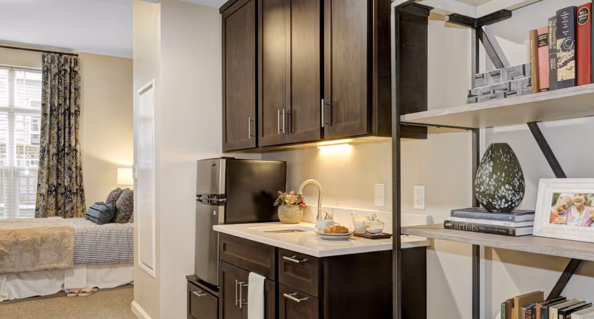 A studio apartment kitchenette with dark wood cabinets, a mini refrigerator and sink opens onto a bed and windowed sleeping area, with shelving holding books and decor.