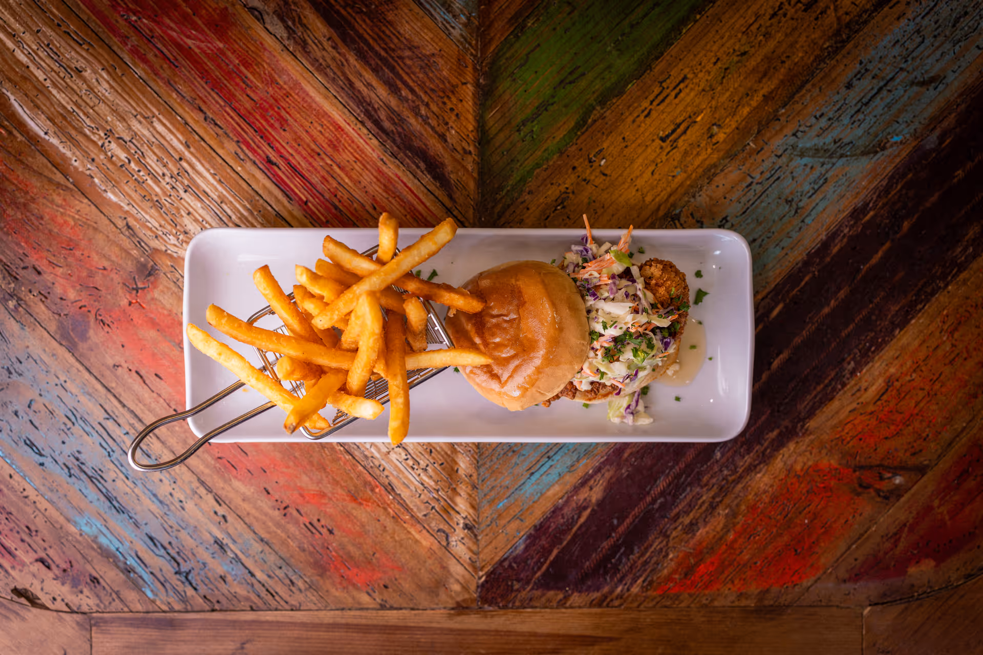 A rectangular white plate on a colorful wooden table holds a serving of French fries in a small metal basket and a sandwich with coleslaw on the side.