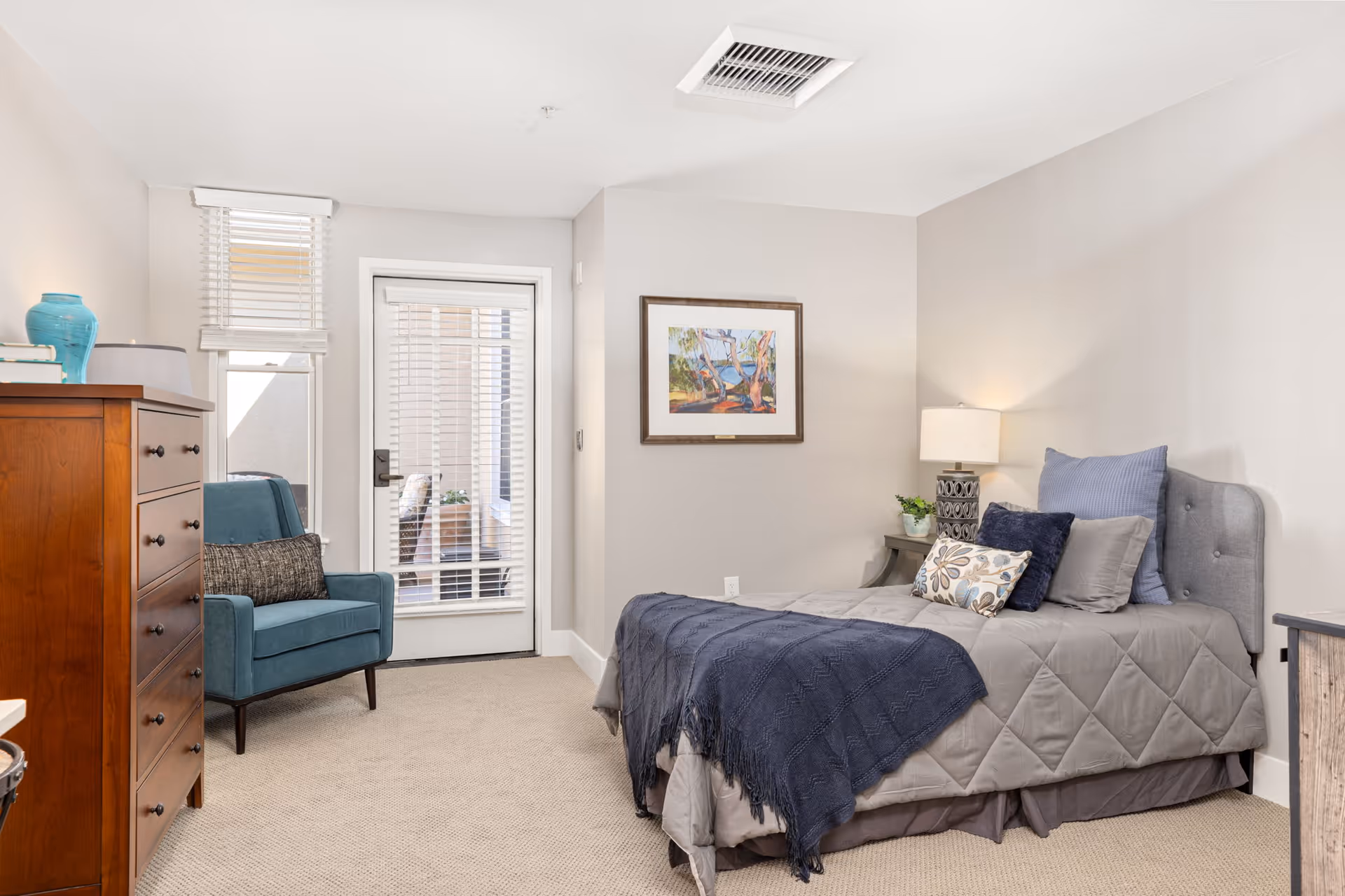 A cozy bedroom with a single bed covered in gray bedding and multiple pillows, a wooden dresser with a blue vase and books on top, a blue armchair with a cushion, a small side table with a lamp and plant, a framed painting on the wall, and a door with blinds leading to an outdoor area.
