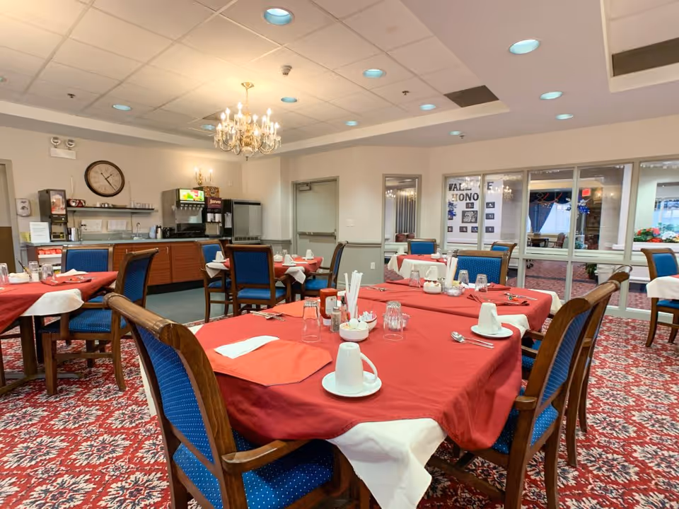 A dining room with several tables covered in red tablecloths and white napkins. Each table is set with cups, glasses, and utensils. Blue cushioned wooden chairs surround the tables. The room has a patterned red carpet, a chandelier hanging from the ceiling, and a beverage station with coffee machines and a clock on the wall in the background. Large windows and a door are visible on one side of the room.