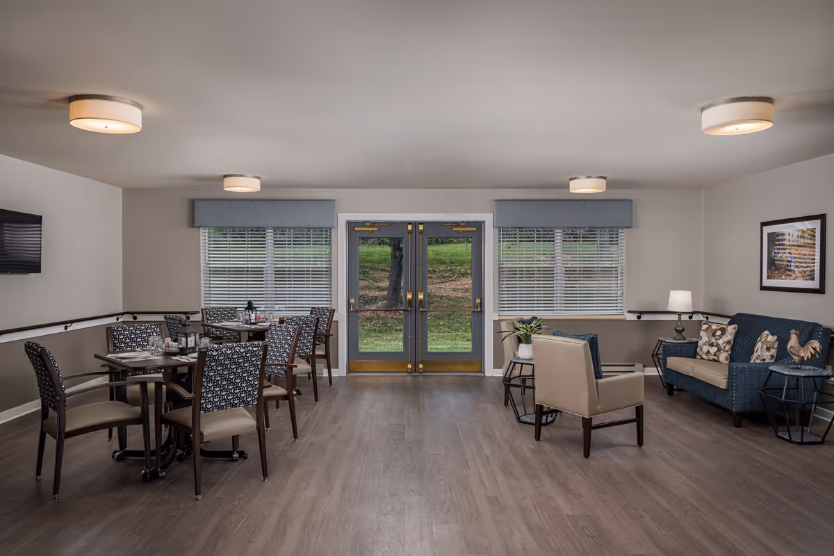 Interior view of a senior living facility common area with a seating arrangement including a blue couch, beige armchair, and side tables with lamps and decorative items. There are two windows with blinds and a double glass door leading outside. On the left side, there are dining tables with patterned chairs and place settings. The room has wood flooring and ceiling lights.