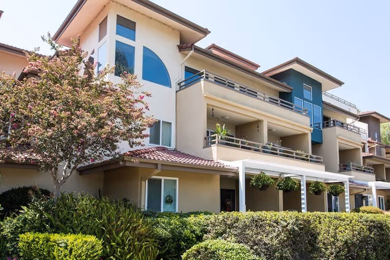 Exterior view of a multi-story residential building with balconies, large windows, and a tiled roof. The building is surrounded by well-maintained greenery, including bushes and a flowering tree, under a clear blue sky.