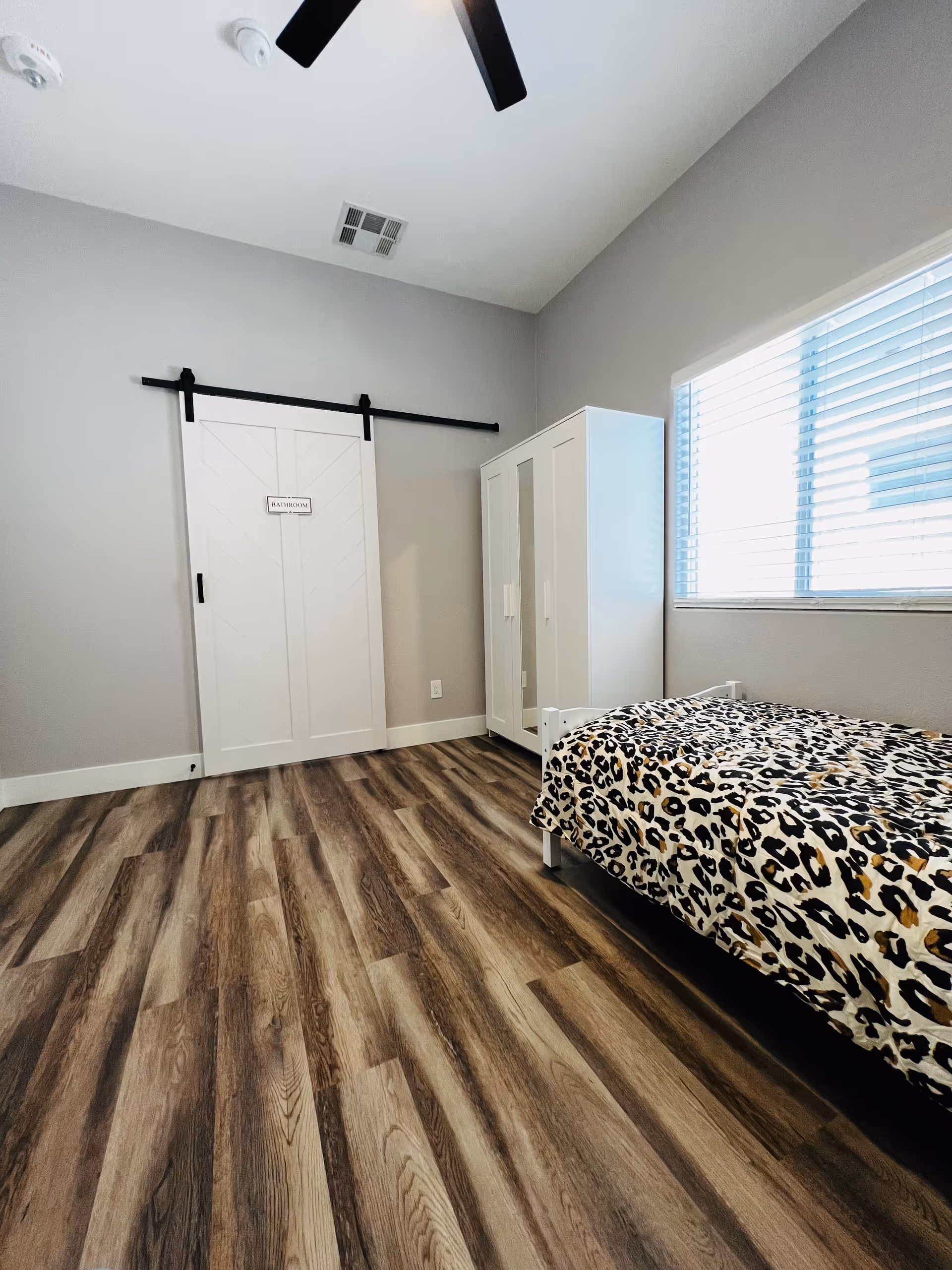 A small bedroom with wood-patterned flooring, a single bed with leopard print bedding, a white wardrobe with mirrored doors, a window with blinds, and a white sliding barn door labeled 'BATHROOM'.