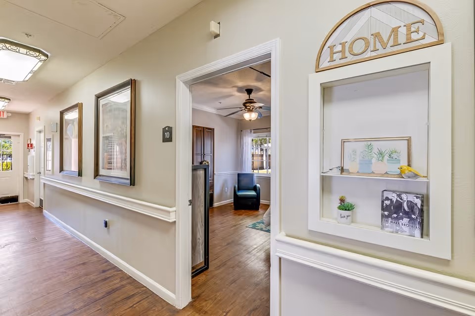 A hallway in a senior living facility with wooden flooring and beige walls. There are framed pictures on the left wall and a small display shelf on the right wall with the word 'HOME' above it. The display shelf contains small decorative items including a picture of The Marx Brothers and small potted plants. An open door reveals a room with a ceiling fan, a blue armchair, and a window with curtains.
