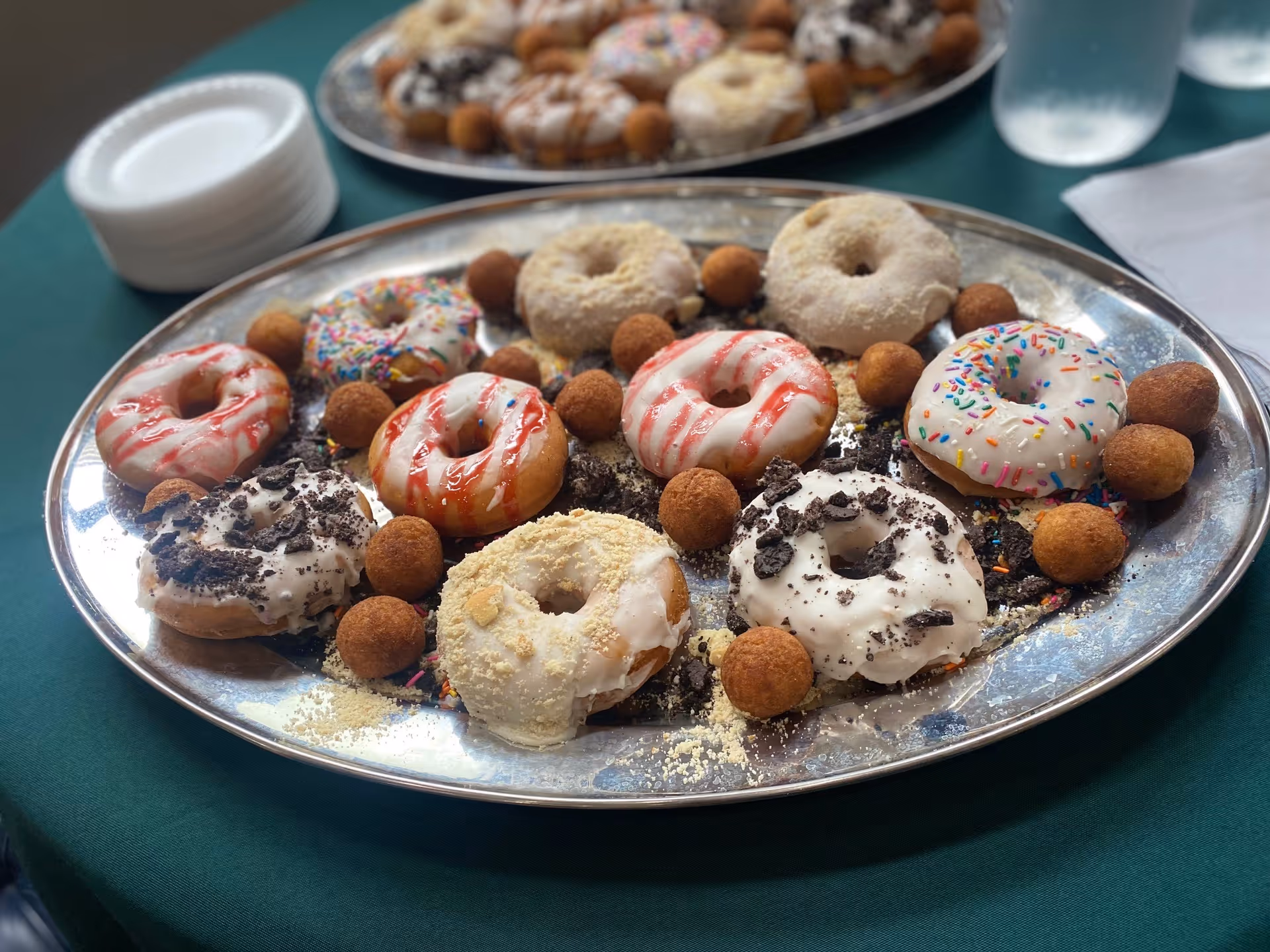 A silver tray filled with assorted decorated donuts and small round donut holes on a green tablecloth. In the background, there is another tray with more donuts, a stack of white plates, a glass of water, and a white napkin.