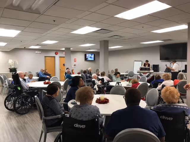 A group of elderly people seated around several round tables in a large room, some in wheelchairs, attentively watching two musicians performing at the front. The room has white walls, a tiled ceiling with fluorescent lights, and a large TV mounted on the wall.