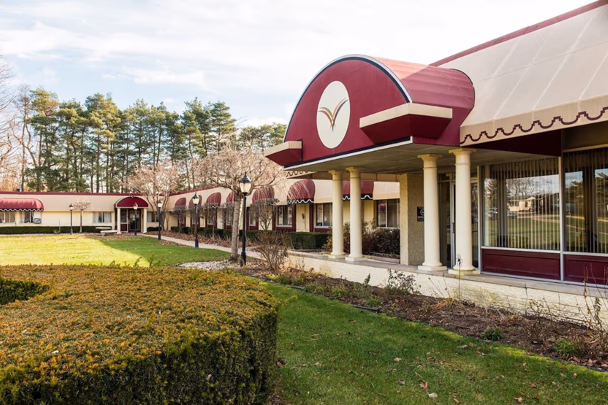 Exterior view of a single-story building with maroon and beige awnings, columns at the entrance, and a well-maintained lawn with trimmed bushes and trees in the background under a partly cloudy sky.