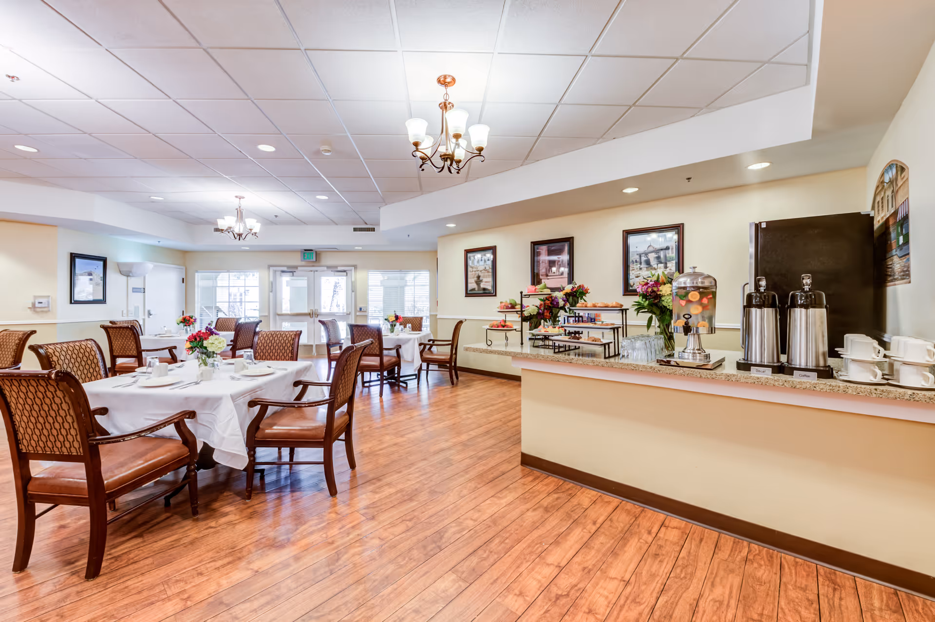 A bright dining room in a senior living facility with wooden floors, several tables covered with white tablecloths, and chairs arranged around them. On the right side, there is a counter with coffee dispensers, a water dispenser with fruit, cups, and a variety of pastries and snacks. The walls are decorated with framed pictures and floral arrangements. Large windows and glass doors allow natural light to enter the room.