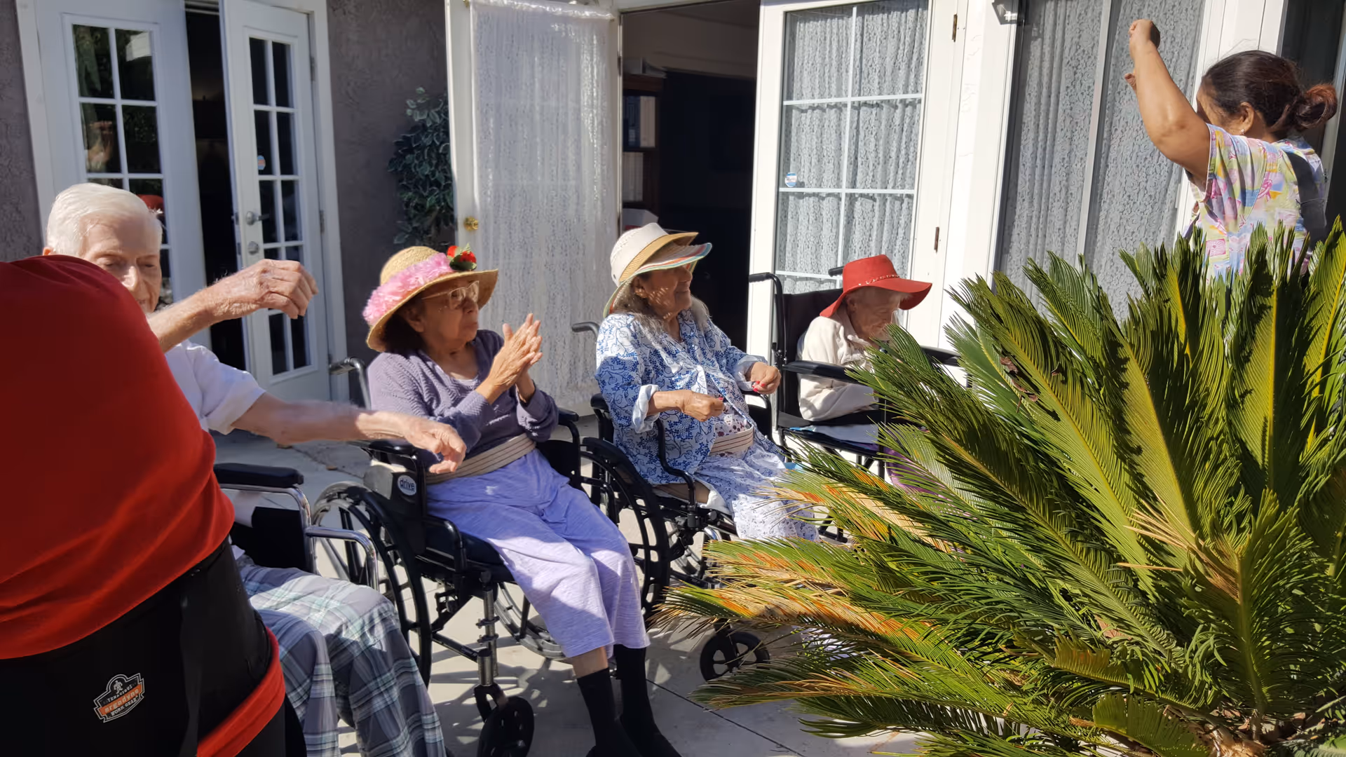 A group of elderly people sitting outside on a patio in wheelchairs, wearing sun hats and enjoying the sunny weather. A caregiver is standing nearby with her arm raised. The background shows glass doors and windows with lace curtains.