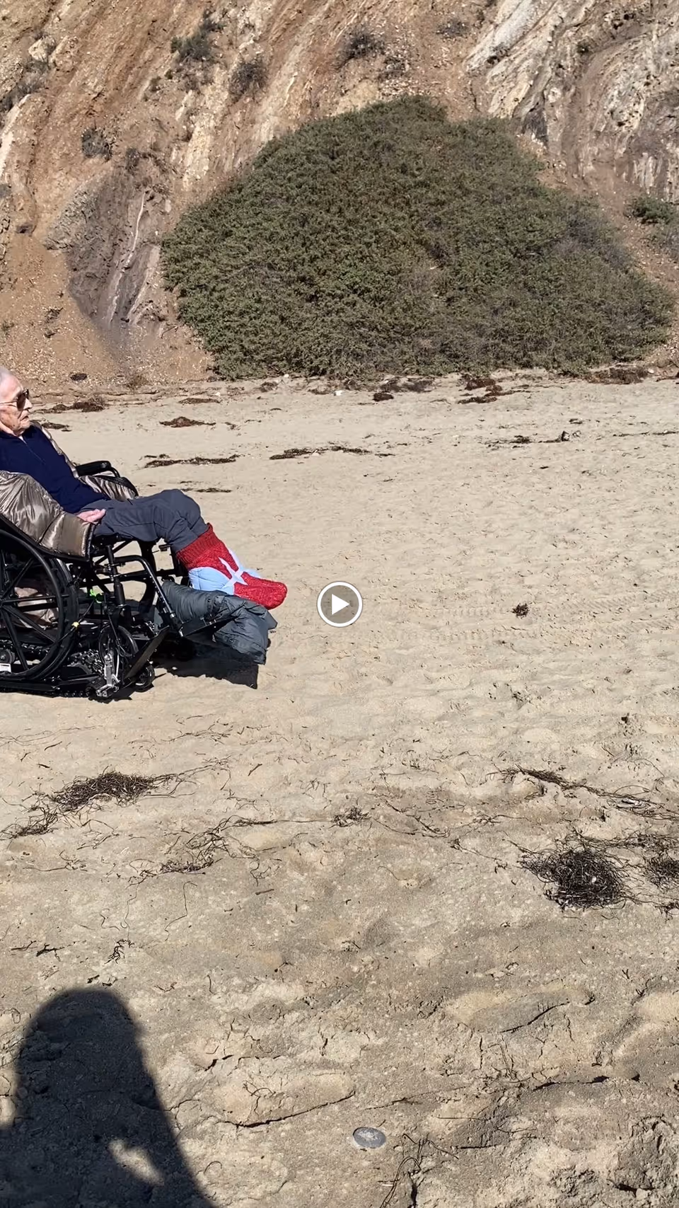 An elderly person sitting in a wheelchair on a sandy beach near a rocky hillside with some vegetation.