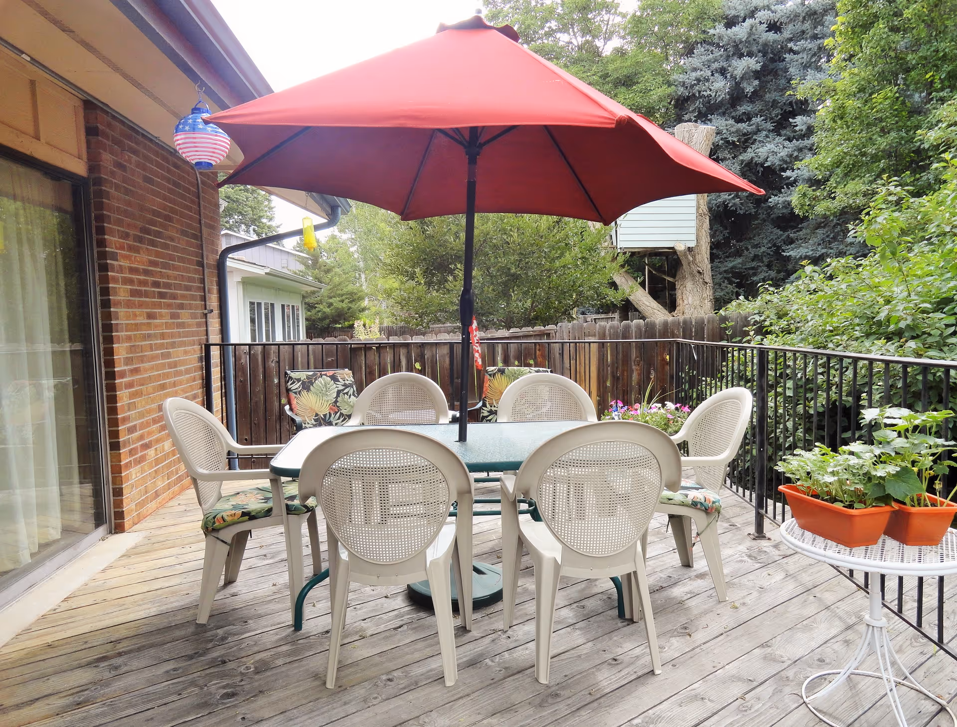 Outdoor wooden deck with a glass-top table surrounded by six white plastic chairs, some with floral cushions. A large red patio umbrella is centered over the table. The deck is bordered by a black metal railing and has potted plants on a small white round table. There are trees and greenery in the background, along with a wooden fence and a brick wall of the building.
