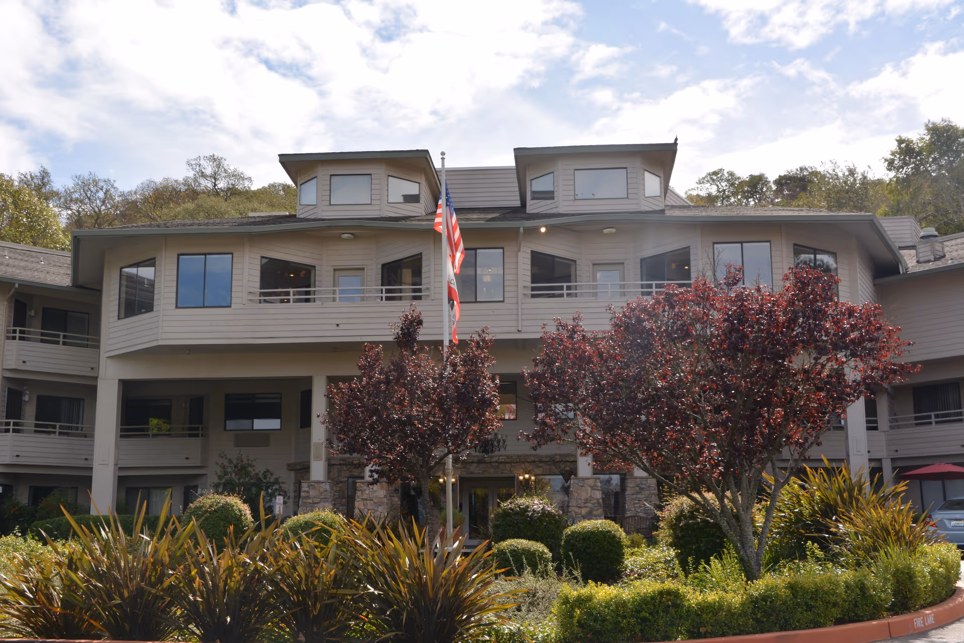 Exterior view of a multi-story retirement community building with beige siding and large windows. The entrance is surrounded by well-maintained landscaping including bushes, red-leafed trees, and other greenery. An American flag and a California state flag are displayed on a flagpole in front of the building under a partly cloudy sky.