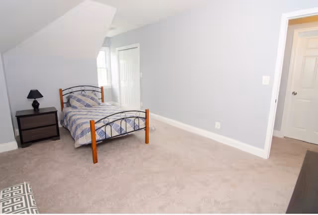 A simple bedroom with a single bed featuring a metal and wood frame, a nightstand with a small lamp, a closet with double doors, and light gray walls with beige carpet flooring.