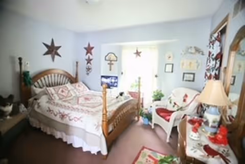 A cozy bedroom with a wooden bed frame, floral quilt, and pillows. The room has light blue walls decorated with star-shaped wall hangings and framed pictures. There is a white armchair with a red cushion, a wooden dresser with a lamp and decorative items, and a window letting in natural light. A cat is sitting on a bench beside the bed.
