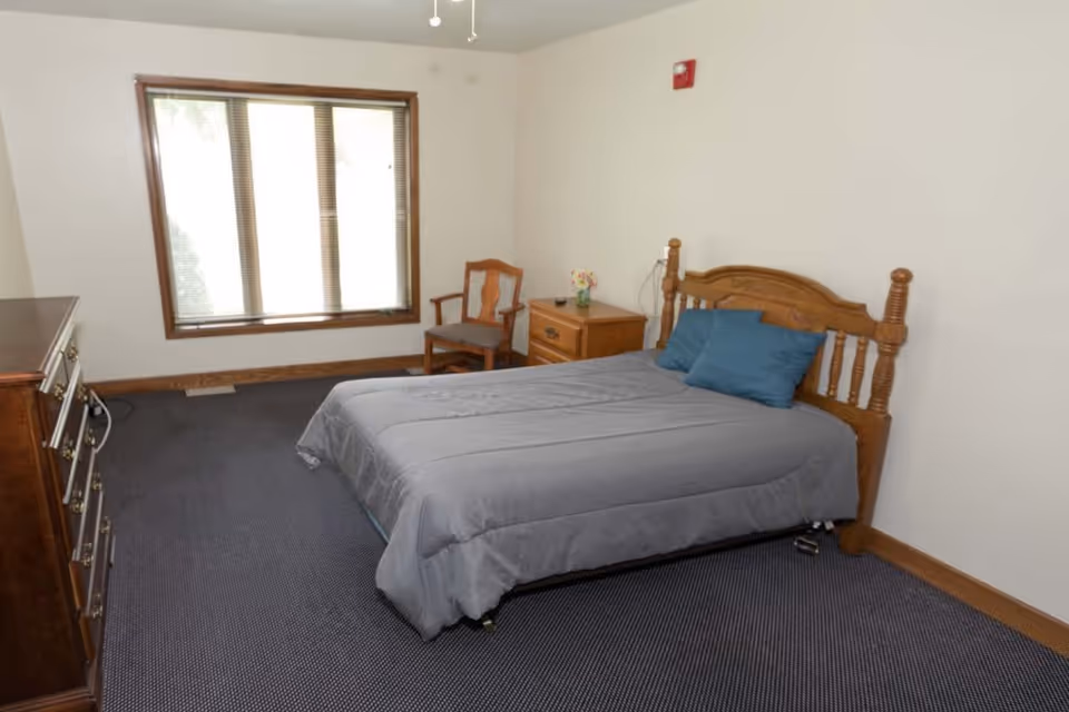 Furnished bedroom with a double bed against the right wall, wooden headboard, dresser and chair, and a large window.