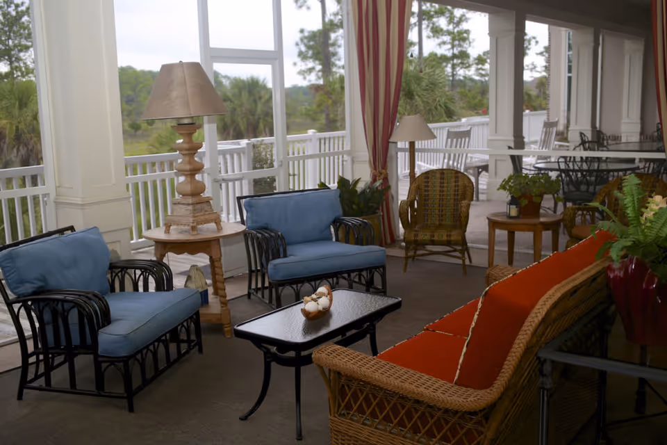 A covered outdoor seating area with wicker and metal furniture including a sofa with orange cushions, two blue cushioned armchairs, and a wicker chair. There are two side tables with lamps and plants, a black coffee table with decorative items, and a dining table with chairs in the background. The area is surrounded by white railings and overlooks greenery and trees.