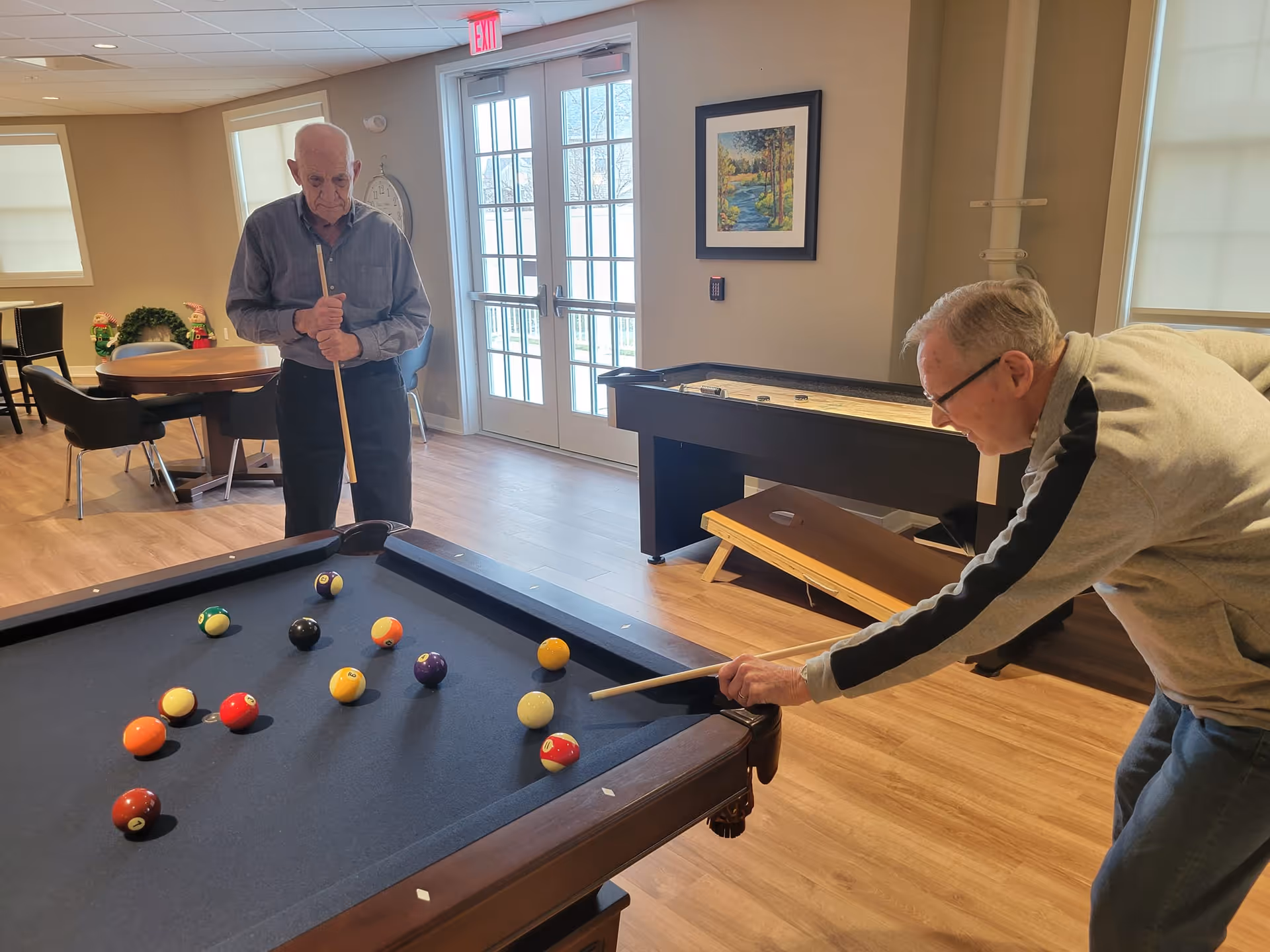 Two men play pool in a bright recreation room with a pool table, shuffleboard, and seating near glass doors.