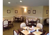Dining room with multiple tables and chairs arranged neatly. Each table is set with placemats, napkins, salt and pepper shakers, and condiments. The walls are light-colored with framed artwork and a wall sconce providing soft lighting.