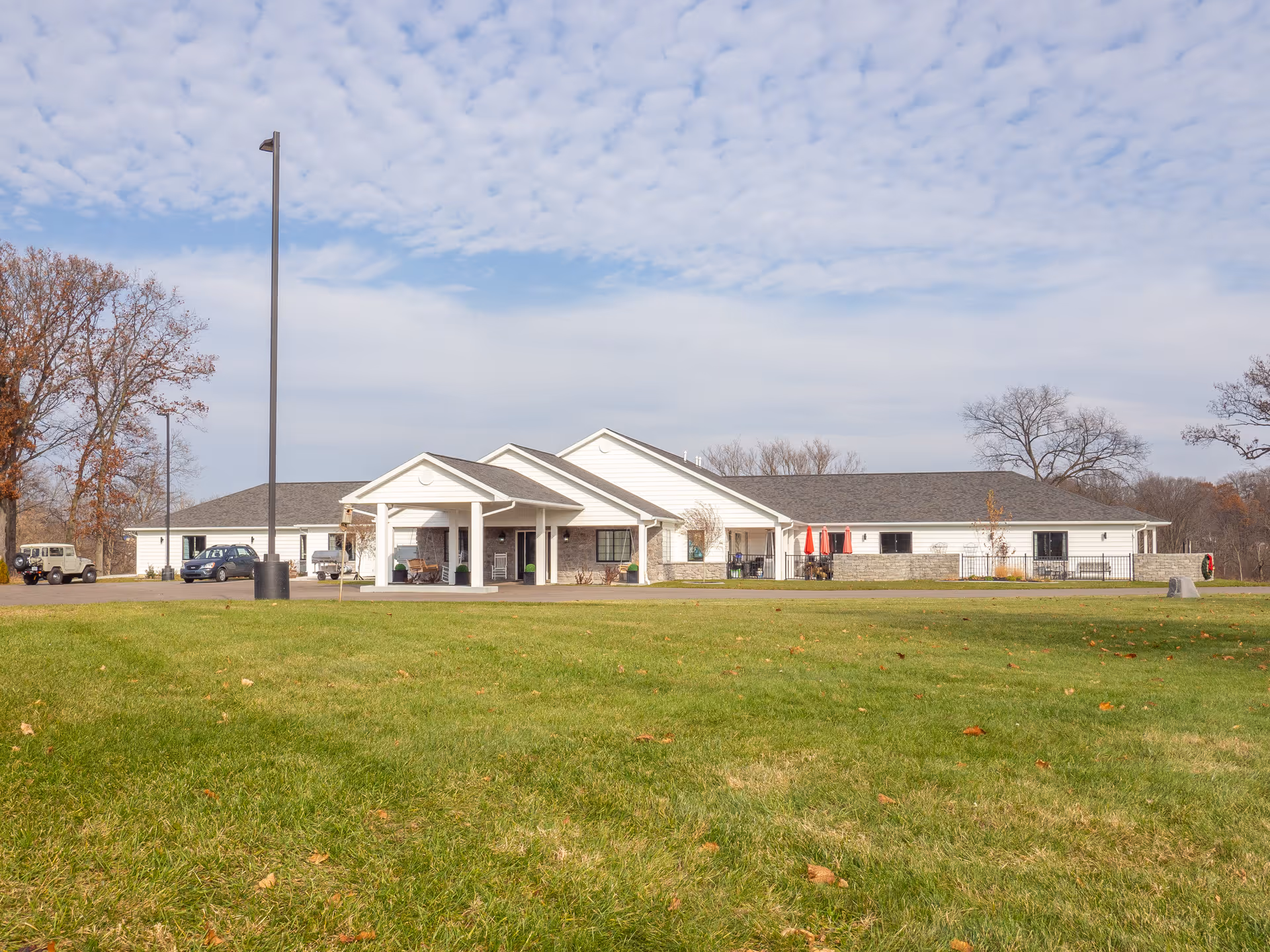 Exterior view of a single-story senior care facility building with white siding and a gray roof, surrounded by a large green lawn and some trees with autumn leaves. There are a few parked cars and a covered entrance with columns.