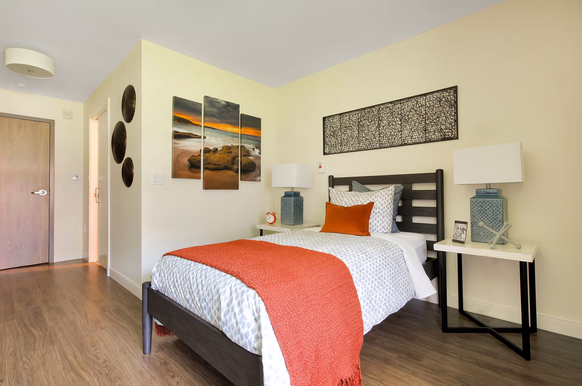 A neatly made single bed with white and gray patterned bedding and an orange throw blanket in a bedroom. There are two bedside tables with blue lamps, a small clock, and decorative items. The walls are light-colored with three round wall hangings and a three-panel beach sunset painting on one wall, and a rectangular abstract artwork above the bed. The floor is wooden, and there is a closed wooden door and an open doorway visible.