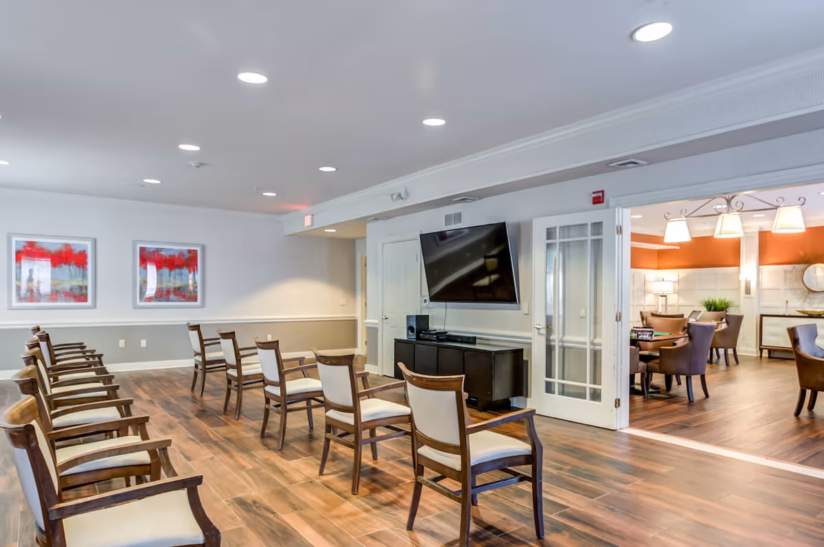 A spacious room with rows of wooden chairs with white cushions facing a large flat-screen TV mounted on the wall above a black cabinet. The room has wood flooring, white walls with a gray lower section, and two framed abstract paintings with red accents. To the right, there is an open doorway leading to another room with brown chairs, tables, and warm lighting.