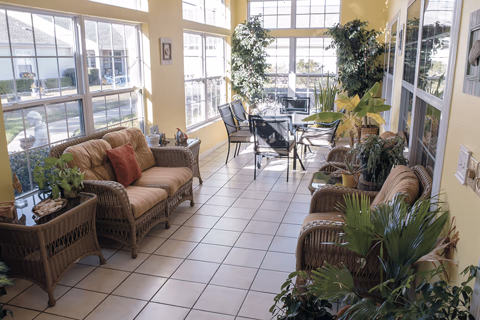 A bright sunroom with large windows on two sides letting in natural light. The room features wicker furniture including two cushioned sofas and a side table with plants. There is a glass-top table with four metal chairs near the windows. Various potted plants are placed around the room, creating a cozy and inviting atmosphere.