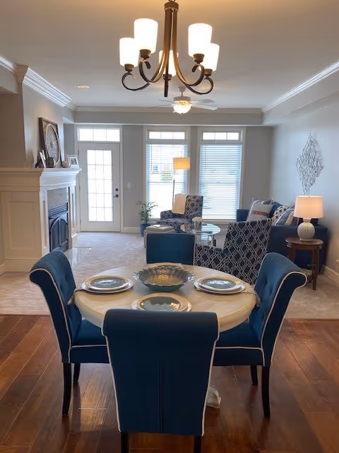 A cozy living and dining area in a senior living facility featuring a round dining table set with four blue upholstered chairs and plates. The living room has two patterned armchairs, a blue sofa, a side table with a lamp, and a fireplace. Large windows and a glass door allow natural light to fill the space.