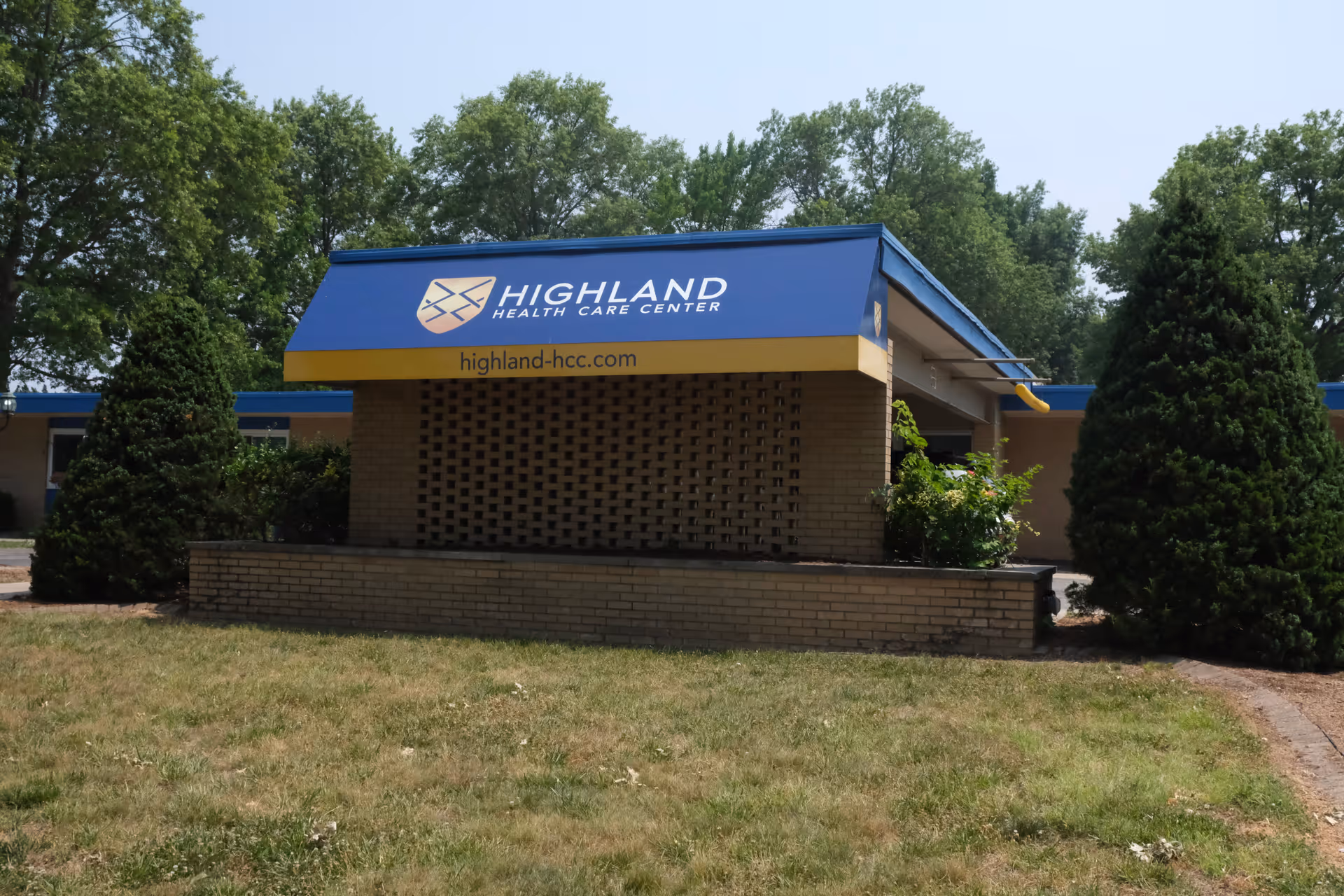 Exterior view of Highland Health Care Center building with a blue and yellow awning displaying the facility's name and website. The building is surrounded by green trees and bushes with a grassy area in the foreground.