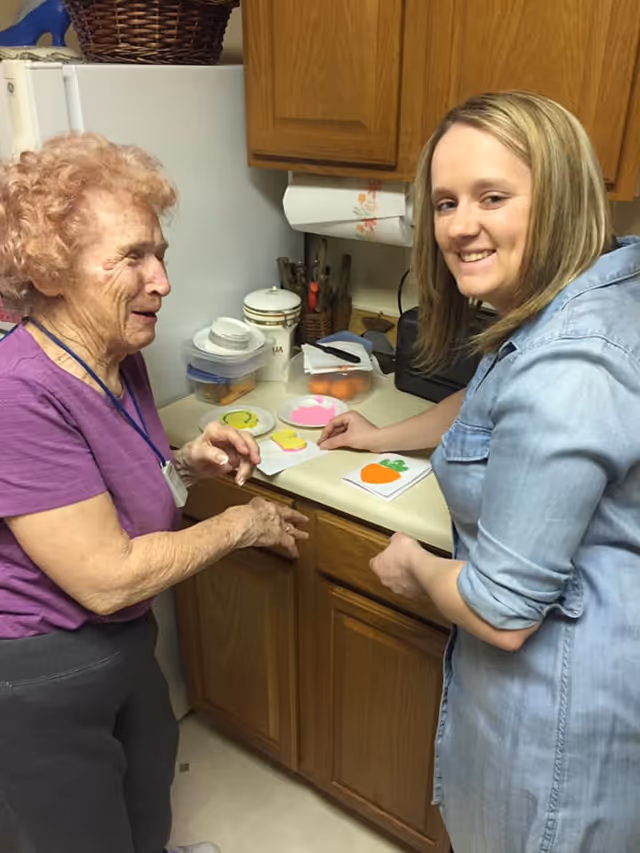 An elderly woman and a younger woman standing in a kitchen. They are smiling and appear to be engaged in an activity involving colorful paper cutouts shaped like a carrot, a duck, and other objects on the kitchen counter. The kitchen has wooden cabinets and various kitchen items on the counter.