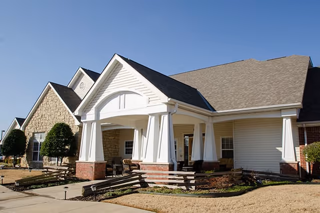 Exterior view of a single-story senior living facility building with a covered entrance supported by white columns, stone and brick accents, and a clear blue sky.