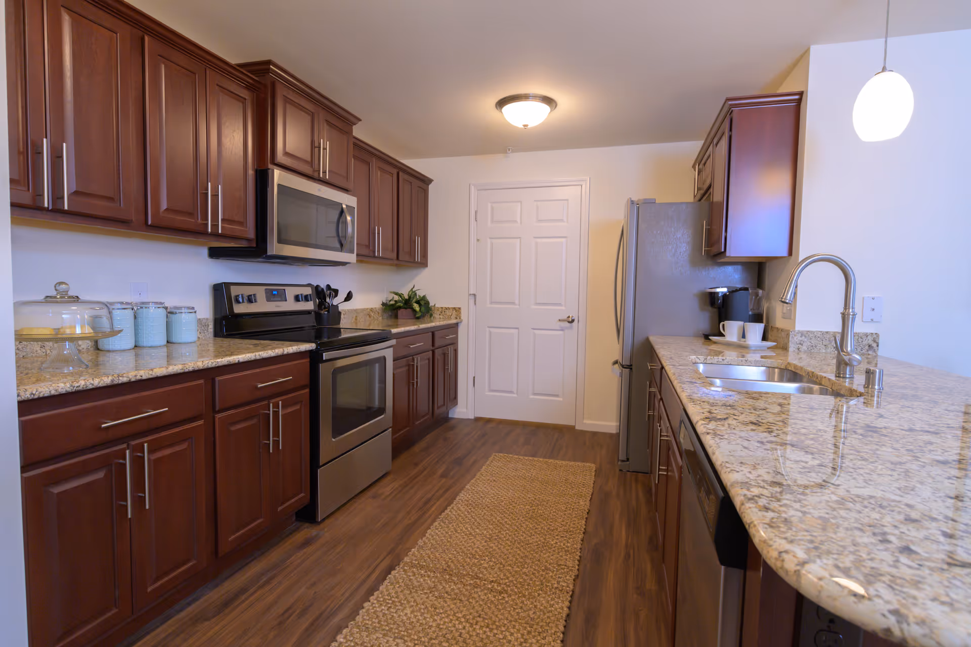 A modern kitchen with wooden cabinets, granite countertops, stainless steel appliances including a stove, microwave, and refrigerator. There is a sink with a curved faucet on the right side, a coffee maker with cups, and a beige woven rug on the wooden floor. The kitchen is well-lit with a ceiling light and a hanging pendant light.