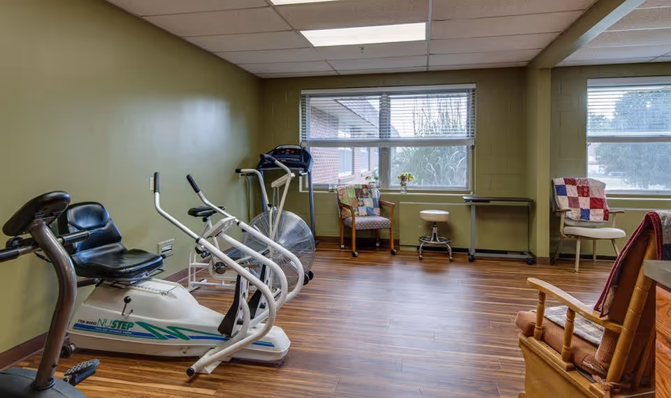 A small exercise room with green walls and wood flooring featuring a recumbent exercise bike, an elliptical machine, and a treadmill. There are several chairs with colorful quilts near two large windows letting in natural light.
