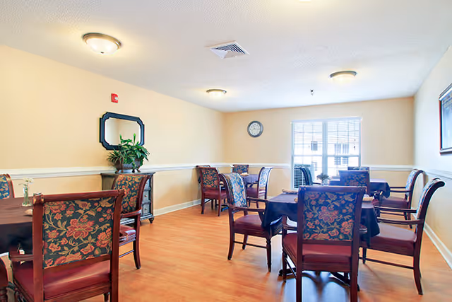 A dining room with several tables covered with dark tablecloths and surrounded by wooden chairs with floral upholstery. The room has light-colored walls, a wooden floor, a wall clock, a window letting in natural light, a mirror, and a small cabinet with a plant on top.