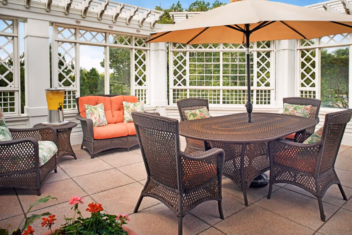 Outdoor patio area with a brown wicker table and five matching chairs, each with floral cushions. There is a large beige umbrella over the table. A loveseat with orange cushions and floral pillows is against a white lattice fence. A beverage dispenser with yellow drink is on a small side table. Green trees are visible through the lattice.
