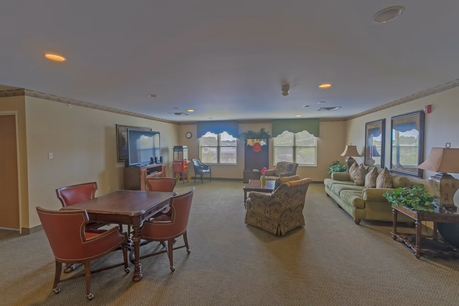 A spacious living room area in a senior living facility with a green sofa, patterned armchairs, a wooden coffee table with a small plant, a wooden table with four red leather chairs, a large flat-screen TV on a wooden stand, two windows with green valances, and decorative plants and lamps on side tables.