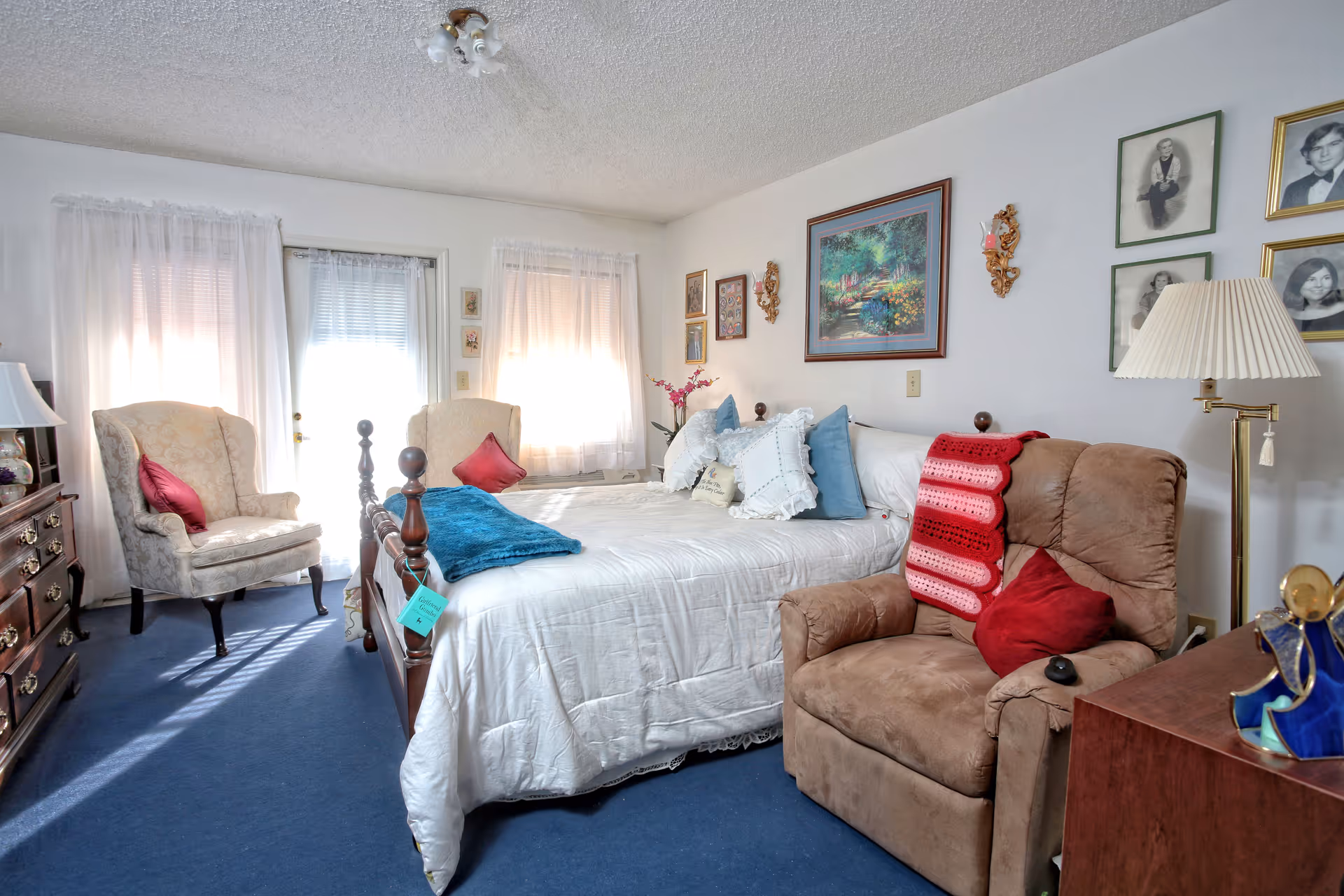 A cozy bedroom with a large bed covered in white bedding and multiple pillows, a brown recliner chair with a red and pink crocheted blanket and a red pillow, two beige armchairs with red pillows near a window with sheer white curtains, a wooden dresser with a lamp, and framed pictures and artwork on the walls.