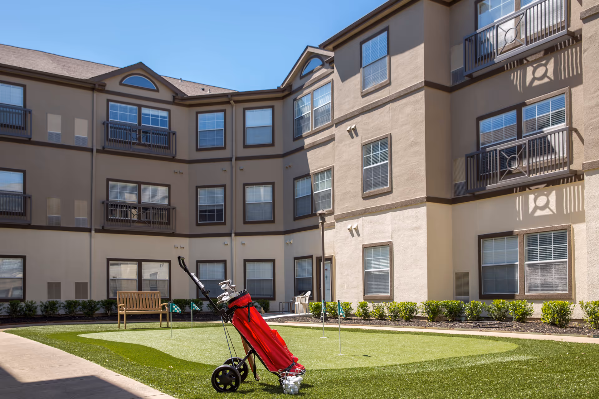 Outdoor courtyard area of a senior living facility with a small putting green, a golf bag on a push cart, a wooden bench, and a three-story building with multiple windows in the background under a clear blue sky.