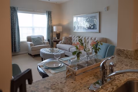 A cozy living room with a beige sofa adorned with patterned pillows, a light blue armchair, and a matching ottoman. A side table with a lamp is next to the sofa. The room has a large window with patterned curtains allowing natural light to fill the space. In the foreground, a granite countertop with a sink and a decorative tray holding glass vases with flowers is visible.