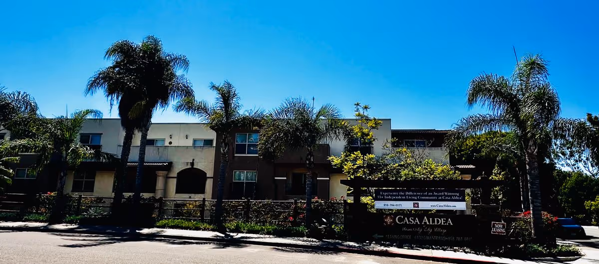 Front exterior of the Casa Aldea senior living building with palm trees and a large entrance sign along the street.