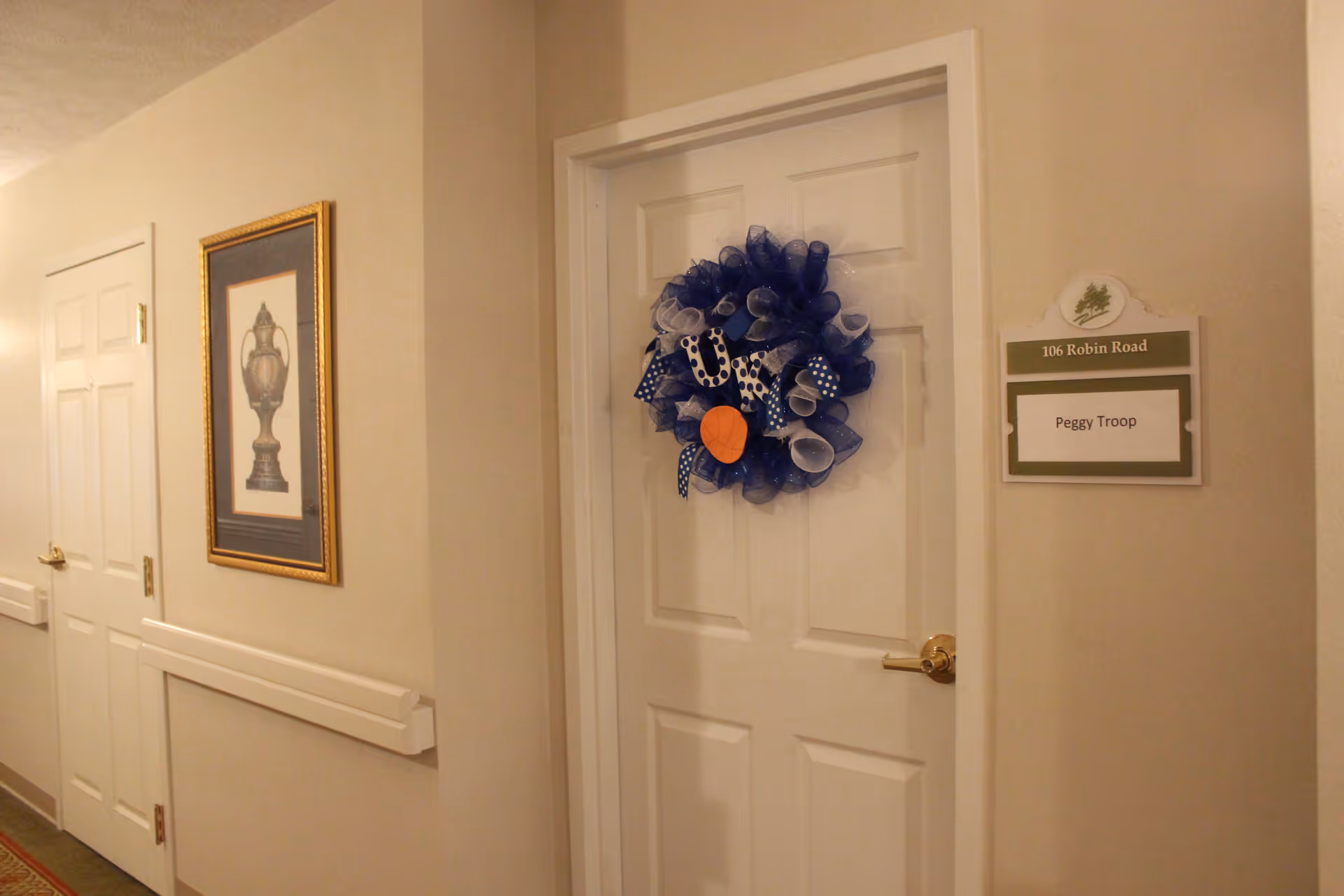 Interior hallway of a senior living facility with beige walls and white doors. One door is decorated with a blue and white wreath and has a nameplate next to it reading '106 Robin Road Peggy Troop'. A framed picture hangs on the wall to the left.