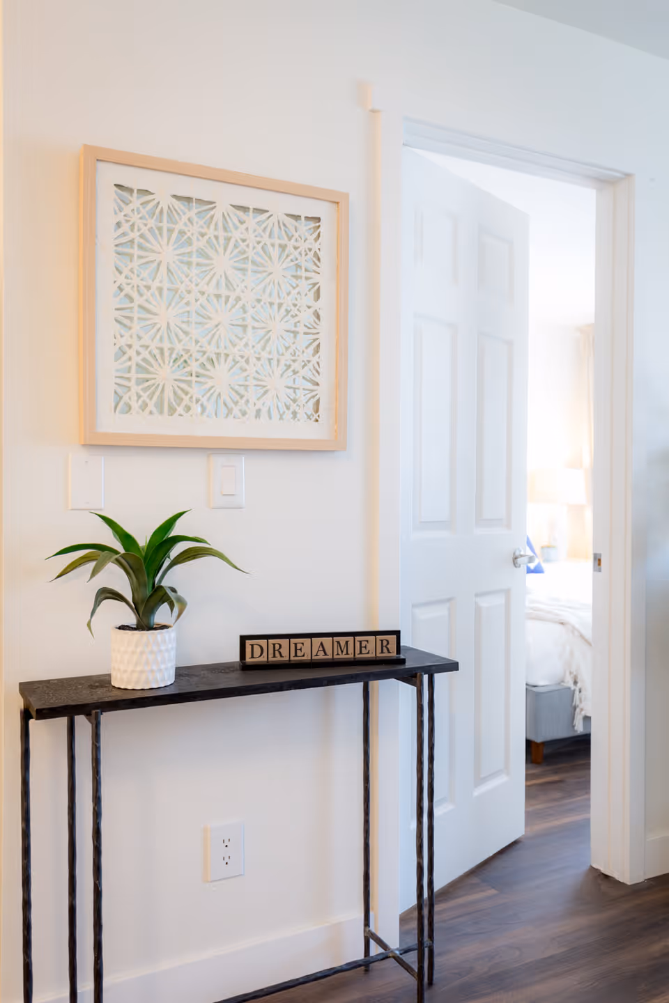 A small black metal console table against a white wall with a potted green plant and wooden blocks spelling 'DREAMER'. Above the table is a framed decorative wall art with a geometric pattern. To the right, a white door is open, revealing a glimpse of a bedroom with a bed, lamp, and wooden floor.
