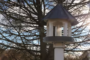 A close-up view of a white bird feeder with a dark roof mounted on a post, set against a backdrop of leafless trees and a clear sky.