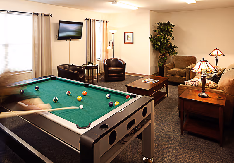 Recreation lounge featuring a pool table in the foreground and a seating area with chairs, sofa, TV, and lamps in the background.