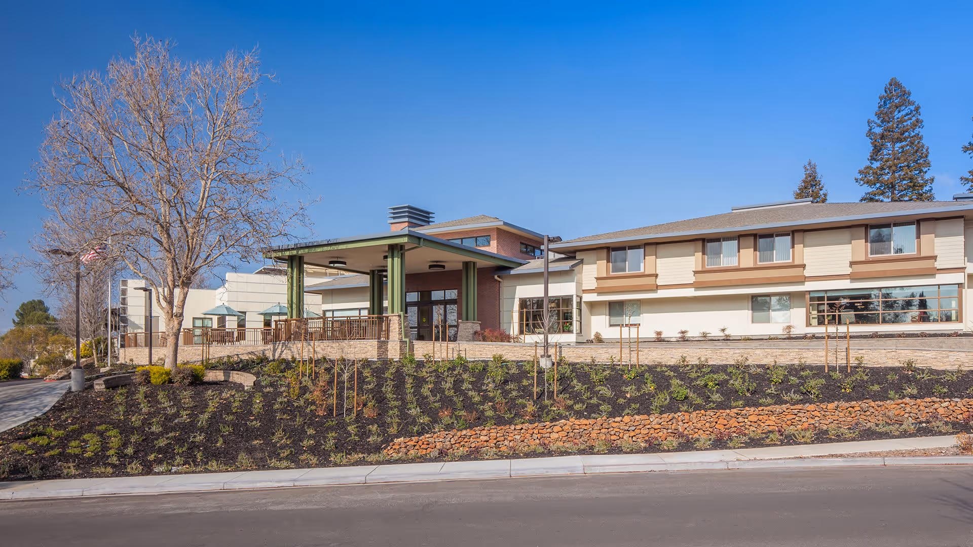 Exterior view of Atria Walnut Creek senior living facility showing a two-story building with large windows, a covered entrance with green pillars, landscaped garden beds, and a clear blue sky.