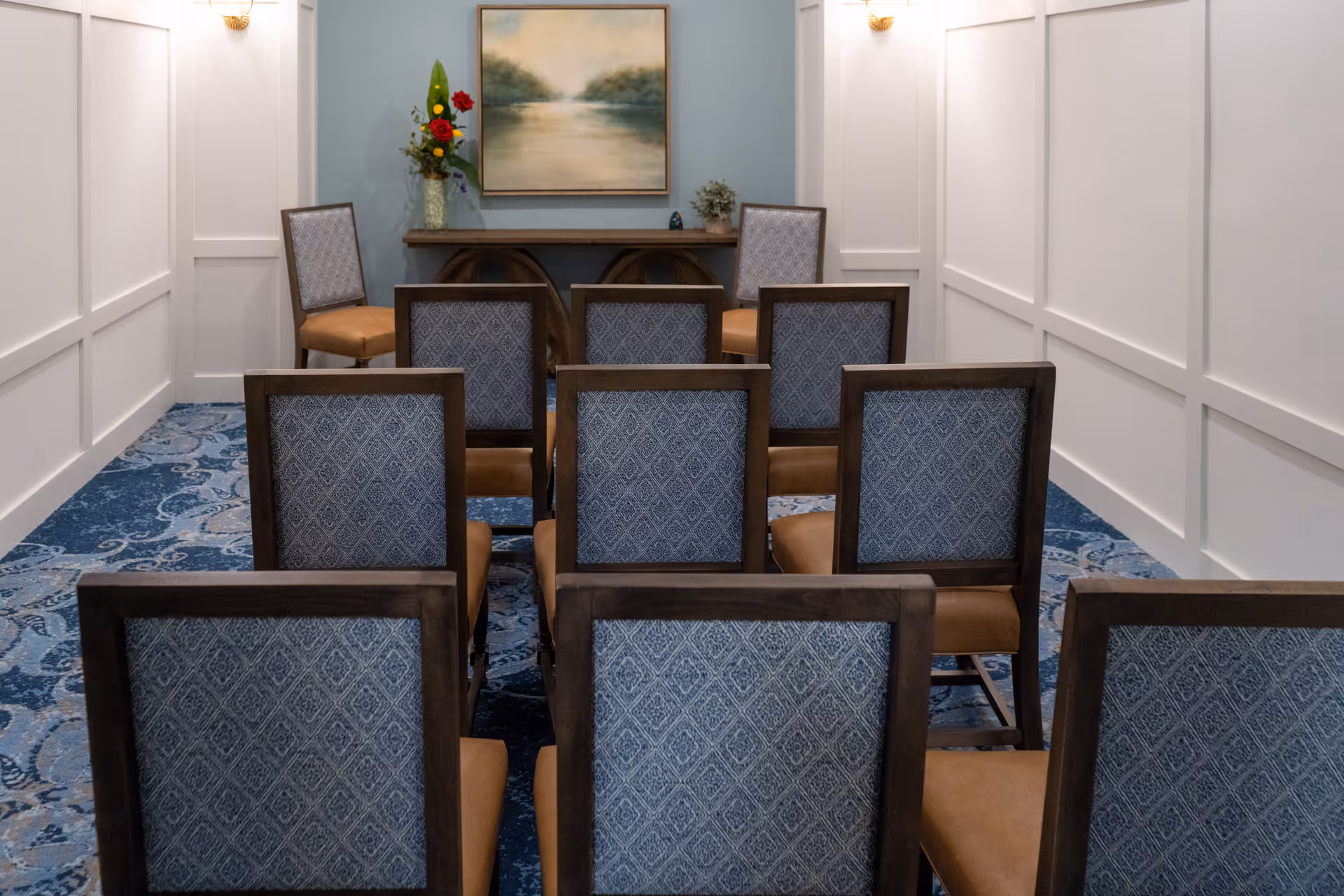 A small meeting or conference room with rows of chairs facing a wooden table against a blue accent wall. On the table is a vase with flowers and a small plant, and above it hangs a framed landscape painting. The room has white paneled walls and a blue patterned carpet.