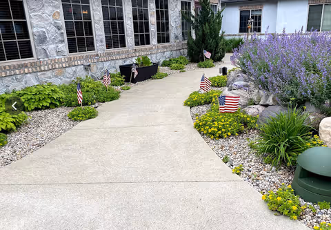 Concrete walkway bordered by rock beds, yellow and purple flowering plants and small American flags leading to a stone-faced building with windows.